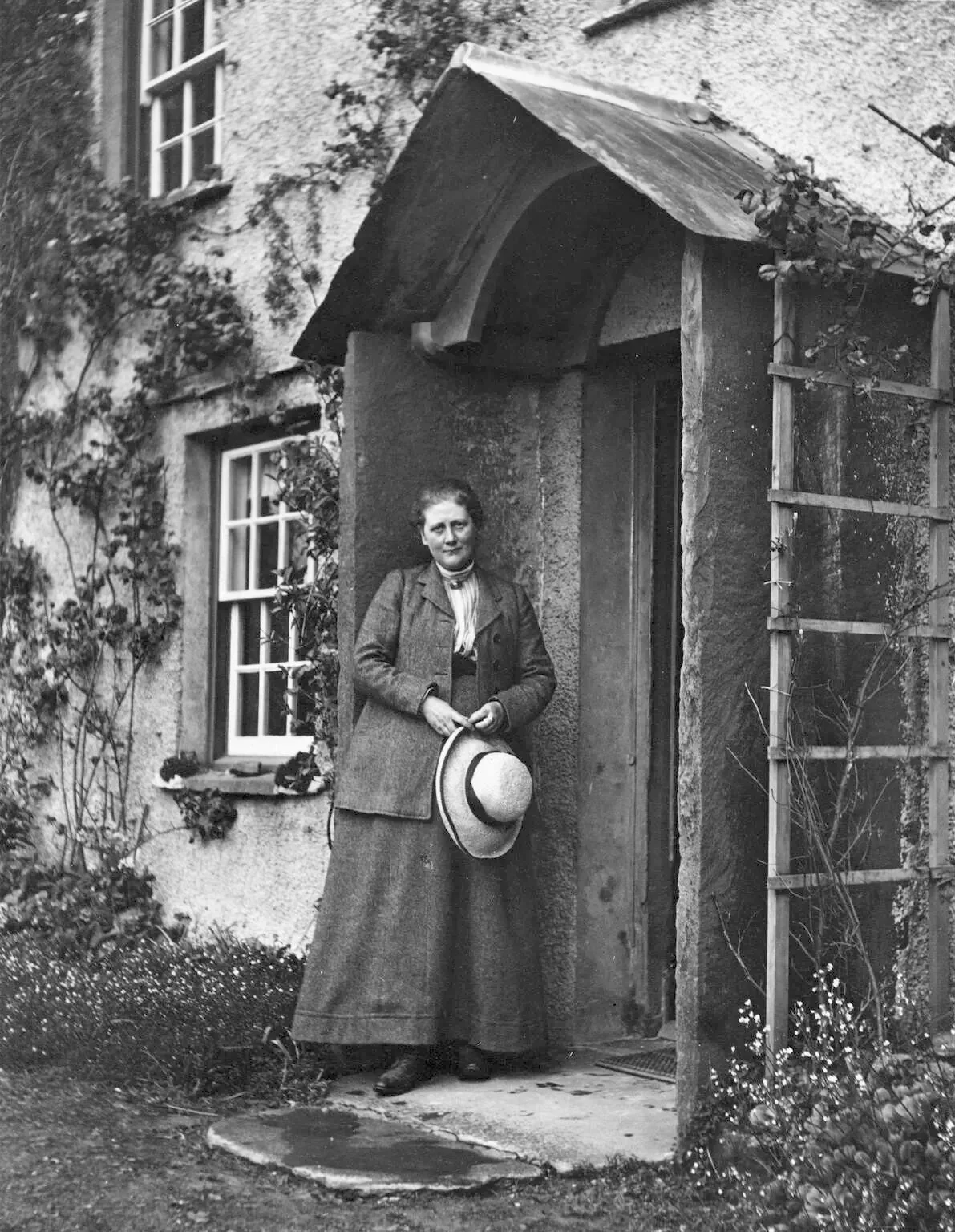 Beatrix Potter standing in a cottage doorway, holding a hat.